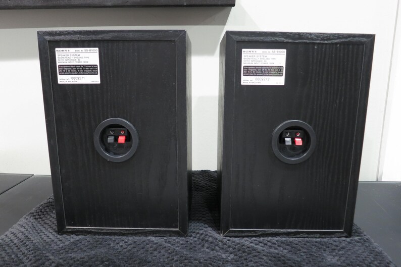 May include: Two black Sony SS-B1000 speaker cabinets shown from the rear. Each unit features a white rectangular label with technical specifications, a circular input terminal housing with one red and one black spring-clip connector, and wood grain texture.