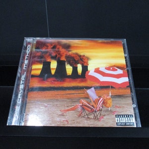 May include: CD case for "The Crystal Method" album. The cover art depicts a beach scene with a person in a beach chair under a red and white umbrella, with smokestacks in the background. The text "THE CRYSTAL METHOD" is visible.
