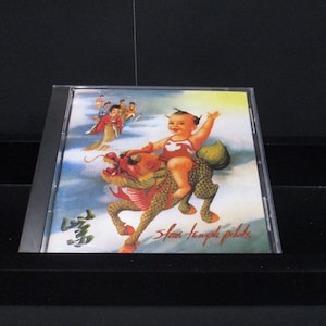 May include: A CD case with a colorful image of a baby riding a dragon. The baby is wearing a red and white outfit and has a crown on its head. The dragon is green and gold and has a red head. The background is a cloudy sky. The text "Slow Temple Pilots" is written in black at the bottom of the image.