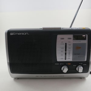 May include: Black and silver AM/FM weather band radio with a digital clock display. The radio has a telescoping antenna and control knobs. The brand name "Emerson" is visible on the front. The radio is designed to receive AM/FM and weather band signals.