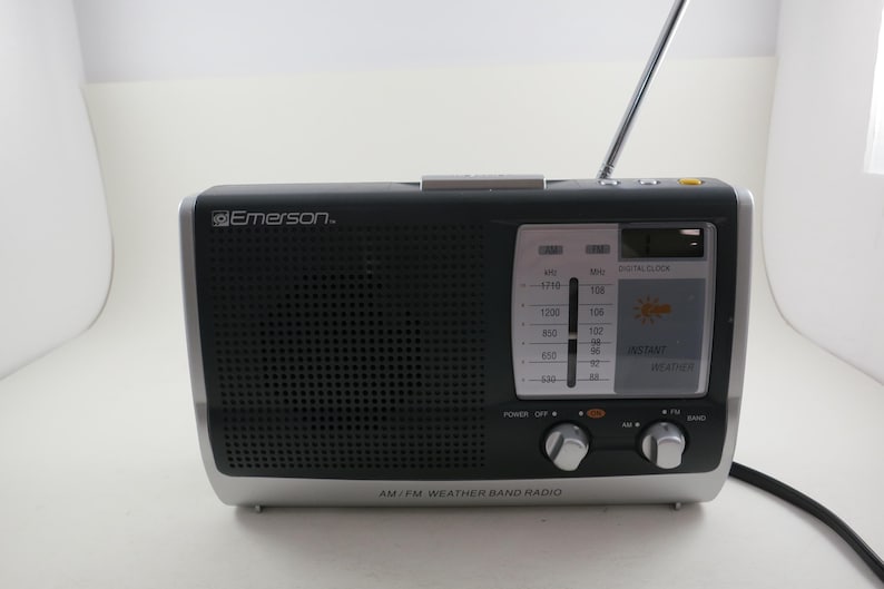 May include: Black and silver AM/FM weather band radio with a digital clock display. The radio has a telescoping antenna and control knobs. The brand name "Emerson" is visible on the front. The radio is designed to receive AM/FM and weather band signals.