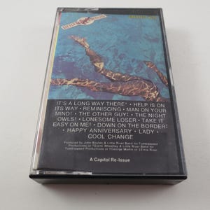 May include: A black cassette tape with a blue and brown image of a person swimming underwater. The cassette tape is labeled "Cool Change" and lists the songs "It's a Long Way There", "Help is on Its Way", "Reminiscing", "Man on Your Mind", "The Other Guy", "The Night Owls", "Lonesome Loser", "Take It Easy on Me", "Down on the Border", "Happy Anniversary", "Lady", and "Cool Change". The cassette tape is a Capitol Re-Issue.