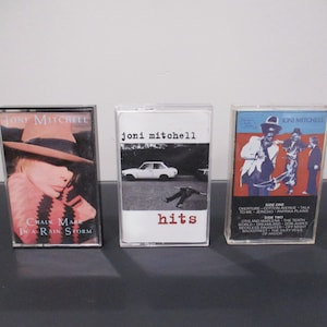 May include: Three vintage cassette tapes featuring Joni Mitchell albums. The covers display album art with text including "Chalk Mark in a Rain Storm" and "hits". The tapes are in clear plastic cases.