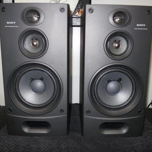 May include: A pair of black Sony speakers. Each speaker has a three-way design, with a large central woofer, a mid-range speaker, and a tweeter. The speakers are rectangular with a small rectangular port at the bottom.
