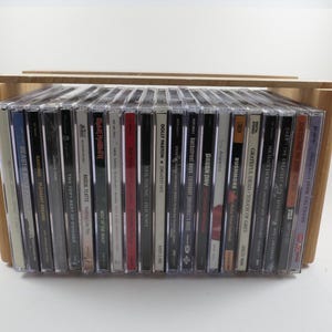 May include: A collection of twenty-two music CDs stored in a light-colored wooden crate. The CDs are arranged vertically, displaying their spines with various artist names and album titles including Beastie Boys, Dolly Parton, and Green Day.