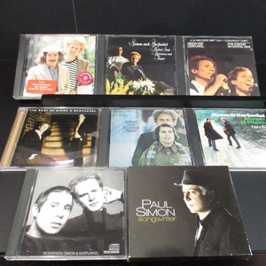 May include: A collection of Simon & Garfunkel and Paul Simon CDs. The album covers feature images of the musicians, album titles, and the text "songwriter". The CDs are displayed on a black shelf.