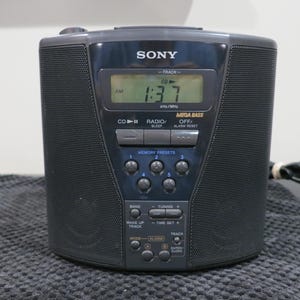 May include: A black Sony portable CD radio alarm clock featuring a digital display showing 1:37 AM, a Mega Bass function, five memory preset buttons, and various control buttons for radio tuning, track selection, and alarm settings with a power cord.