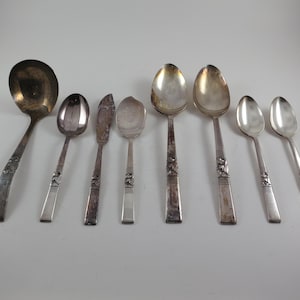 May include: A collection of antique silverware, including spoons and a butter knife, arranged on a white surface. The spoons vary in size and shape, with decorative details on the handles. The metal has a tarnished appearance.
