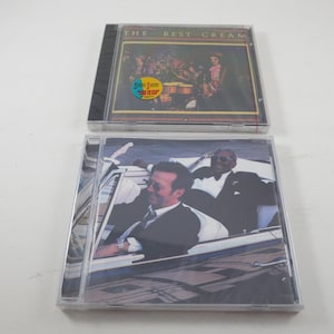 May include: Two music CDs. The top CD case is black with the text "THE BEST CREAM" and a colorful image. The bottom CD case features a black and white image of two men in a car. Both CDs are in clear plastic cases.