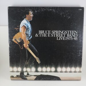 May include: Album cover for Bruce Springsteen & The E Street Band LIVE/1975-85. The cover features a black background with the band's name and album title in white text. A man holding a guitar is walking on stage.