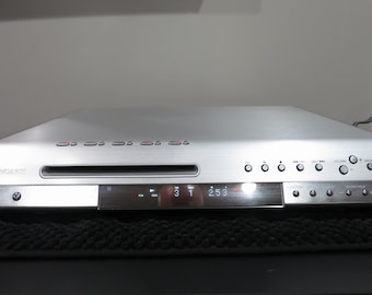 Può includere: Un lettore DVD Sony color argento con una sottile fessura orizzontale per il disco, un display digitale centrale che mostra i numeri 3, 1 e 253 e diversi pulsanti di controllo circolari sul pannello frontale per alimentazione e riproduzione.