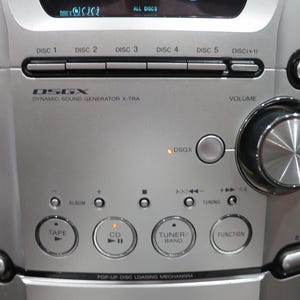 以下が含まれることがあります： 5枚のCDチェンジャー、大きな円形の音量調節ノブ、Tape、CD、Tuner/Band、Functionとラベル付けされたボタンを備えたシルバーのステレオシステム。フロントパネルにはDSGX Dynamic Sound Generator X-Traの文字とEnergy Starのロゴが表示されています。