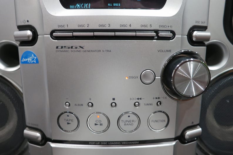 以下が含まれることがあります： 5枚のCDチェンジャー、大きな円形の音量調節ノブ、Tape、CD、Tuner/Band、Functionとラベル付けされたボタンを備えたシルバーのステレオシステム。フロントパネルにはDSGX Dynamic Sound Generator X-Traの文字とEnergy Starのロゴが表示されています。
