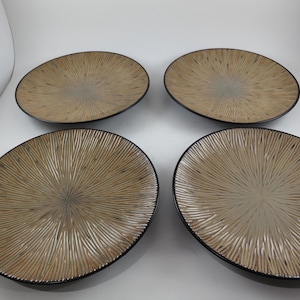May include: Four decorative oval plates. Each plate has a beige center with a radial textured pattern and a black rim. The plates are arranged in a square formation against a white background.