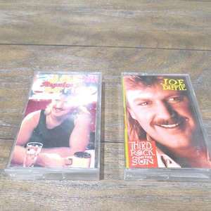 May include: Two vintage cassette tapes featuring country music artist Joe Diffie. One tape is labeled "Regular Joe" and the other "Third Rock From The Sun". Both tapes are in clear plastic cases and are set against a wooden background.