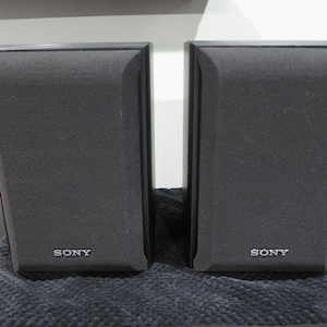 May include: A pair of black Sony bookshelf speakers with rectangular frames and fabric grilles. Each speaker features the Sony brand name printed in white lettering at the bottom center of the front panel. The speakers have a clean, vertical design.