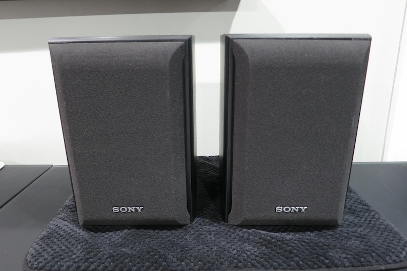 May include: A pair of black Sony bookshelf speakers with rectangular frames and fabric grilles. Each speaker features the Sony brand name printed in white lettering at the bottom center of the front panel. The speakers have a clean, vertical design.