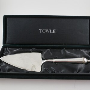 May include: A silver cake server in a black velvet-lined box. The server has a triangular blade and a decorative handle. The box is open, revealing the server resting on black satin fabric. The word "TOWLE" is printed on the inside of the box.