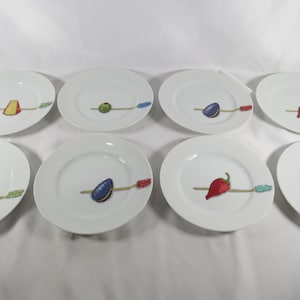 May include: A set of eight white ceramic plates, each featuring a central graphic of a food item skewered on a decorative stick. The food illustrations include cheese, an olive, a plum, a chili pepper, shrimp, and a mussel, all in color.