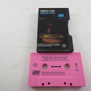 May include: A vintage audio cassette tape and its case. The cassette is pink with the text "ROBERTA FLACK Killing Me Softly" and song titles. The case is black with the album art and text. The cassette is from 1973.