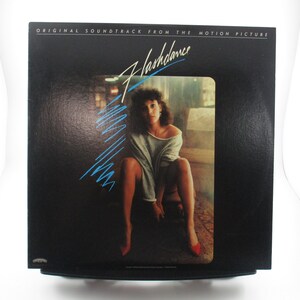 May include: A black vinyl record album cover with the title "Flashdance" in white text. The cover features a woman in a white shirt and jeans, sitting on a ledge with a blue and white abstract design in the background.