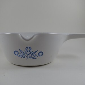 May include: A white ceramic saucepan with a long handle and a pouring spout on the side. The exterior features a blue floral pattern consisting of small flowers and leaves centered on the side of the bowl, providing a simple and clean aesthetic.