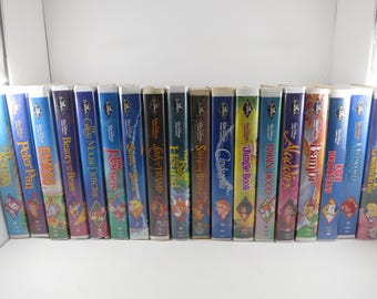 May include: A collection of seventeen Walt Disney VHS tapes arranged in a row, featuring titles such as Peter Pan, Beauty and the Beast, The Rescuers, Lady and the Tramp, The Fox and the Hound, Sleeping Beauty, Cinderella, The Jungle Book, and Aladdin.