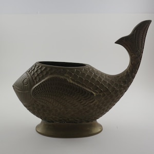 May include: A bronze-colored fish-shaped decorative vase. The vase has a detailed scale pattern covering its body and a curved tail. The vase sits on a round base and has an open top for holding items. The fish's eye is visible on the side.