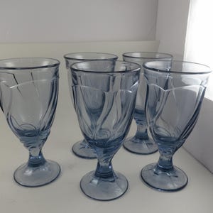 Pu&ograve; includere: Un set di cinque calici in vetro colorato di blu. Ogni calice presenta una coppa a forma di tulipano con un design decorativo a spirale e una base arrotondata. I bicchieri sono esposti su una superficie bianca.