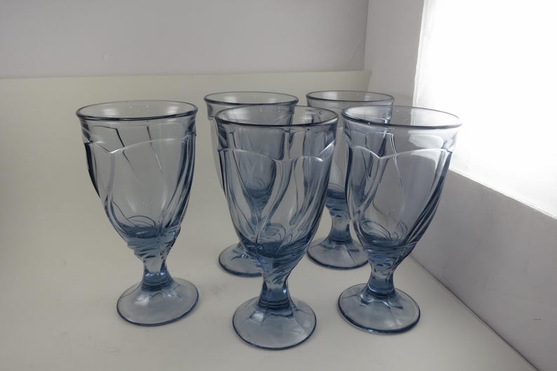 Pu&ograve; includere: Un set di cinque calici in vetro colorato di blu. Ogni calice presenta una coppa a forma di tulipano con un design decorativo a spirale e una base arrotondata. I bicchieri sono esposti su una superficie bianca.