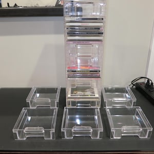 May include: A clear, plastic CD storage tower with multiple compartments, holding several CDs. Six matching clear plastic trays are arranged in front of the tower. The items are on a dark surface.