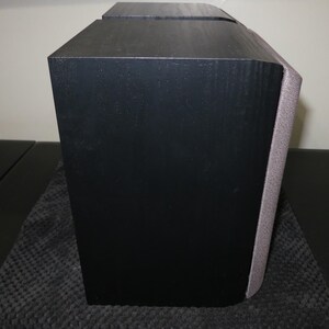 May include: A black speaker with a textured finish. The speaker has a rectangular shape with a curved edge on one side. A gray fabric covers the curved edge. The speaker is on a dark surface.