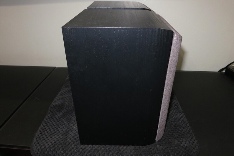 May include: A black speaker with a textured finish. The speaker has a rectangular shape with a curved edge on one side. A gray fabric covers the curved edge. The speaker is on a dark surface.