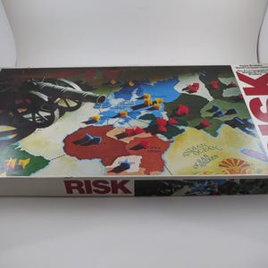 May include: A board game box for Risk, featuring a world map with territories marked by colored pieces. The box has the word "RISK" in large red letters and a cannon illustration.