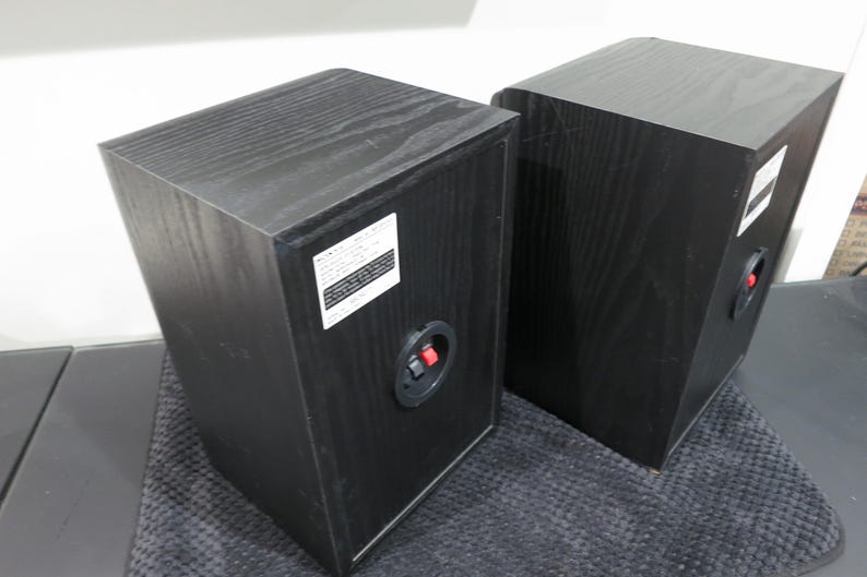May include: Two black wooden bookshelf speakers shown from the rear angle. Each speaker features a rectangular white product information label and a circular connection terminal with one red and one black spring-loaded clip for audio wire input.