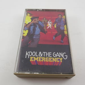 May include: A cassette tape with a black background and a colorful image of a band. The text "Kool & The Gang" and "Emergency" is printed in white on the tape.