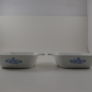 May include: Two white rectangular casserole dishes with blue floral designs. Each dish has a handle on either side. The floral design is a cluster of blue flowers and leaves. The dishes are on a white surface.