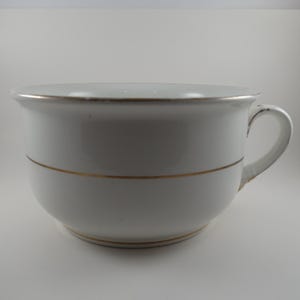 May include: White ceramic bowl with a handle, featuring a gold band around the middle and rim. The bowl has a rounded shape and a handle on the side. The bowl is likely for serving or display.