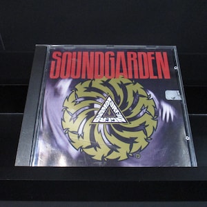 May include: A CD case with a black background and a silver border. The front of the case features a black and white image of a swirling design with a yellow triangle in the center. The text "SOUNDGARDEN" is printed in red letters at the top of the case.
