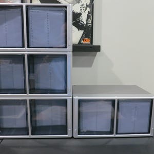 May include: A silver modular storage unit with eight clear-fronted drawers arranged in a vertical stack of six and a horizontal pair of two. Each drawer features the brand name HIPCE printed in the bottom corner and a light gray interior lining.