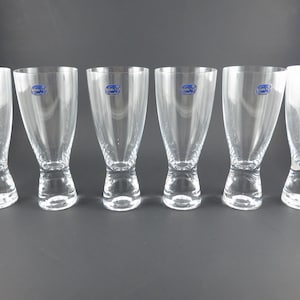 May include: A set of six clear glass tumblers arranged in a row. Each glass features a thick, weighted base that tapers upward into a wider, smooth rim. A small, circular blue sticker with white text is affixed to the upper side of each glass.