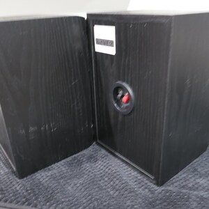 May include: Two black wooden bookshelf speakers with a visible wood grain texture. Each speaker features a circular rear connection port with one red and one black terminal, along with a white rectangular label positioned near the top edge.