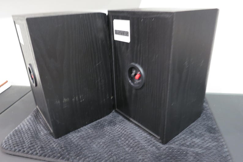 May include: Two black wooden bookshelf speakers with a visible wood grain texture. Each speaker features a circular rear connection port with one red and one black terminal, along with a white rectangular label positioned near the top edge.