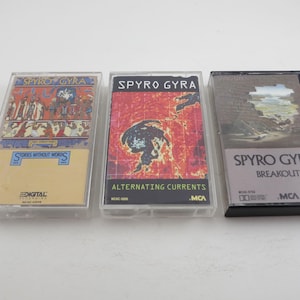 May include: Three cassette tapes by the band Spyro Gyra. The tapes are in clear plastic cases with the band's name and album titles printed on the front. The album titles are "Stories Without Words", "Alternating Currents", and "Breakout".