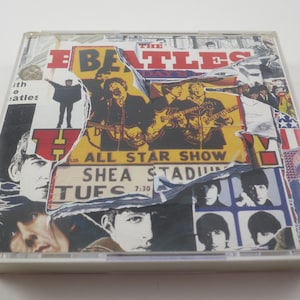 May include: A CD case featuring a collage design with the band name "The Beatles" in red and yellow. The collage includes images of the band members, concert details, and a torn paper effect. The text "ALL STAR SHOW SHEA STADIUM TUES 7:30" is visible.