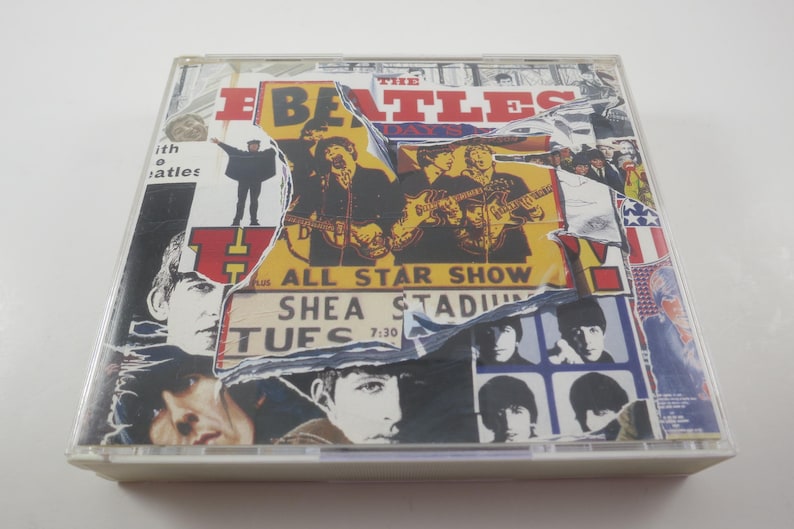 May include: A CD case featuring a collage design with the band name "The Beatles" in red and yellow. The collage includes images of the band members, concert details, and a torn paper effect. The text "ALL STAR SHOW SHEA STADIUM TUES 7:30" is visible.