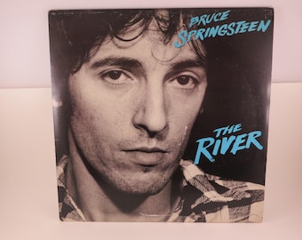 May include: Album cover featuring a black and white portrait of Bruce Springsteen. The text "Bruce Springsteen" is in turquoise script at the top, with "The River" in turquoise script below. The album cover is square.