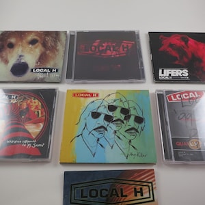May include: Collection of Local H CDs. Album covers feature a dog, abstract art, and band members. Titles include "Lifers" and "Hey Killer". The CDs are in clear plastic cases, with the band's logo and album titles.