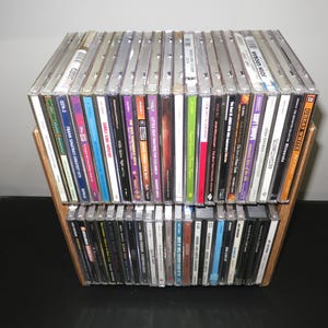 May include: A wooden CD rack filled with compact discs. The rack is constructed with two shelves and angled side supports. The CDs have various colorful album covers and spines, creating a visually diverse collection.