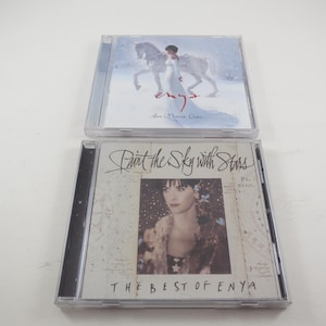 May include: Two compact discs, one with a white background and a woman in a white dress riding a white horse, the other with a white background and a woman with long dark hair wearing a floral dress, the text "Paint the Sky with Stars The Best of Enya" is printed on the front.